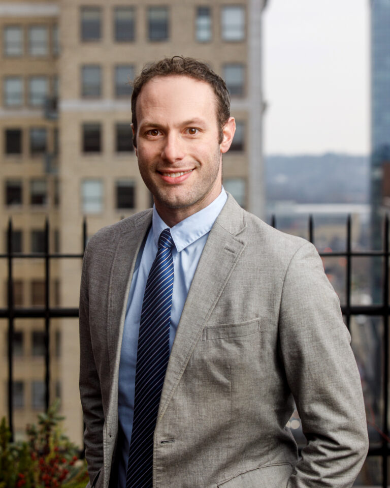 A headshot of Chris Romero, wearing a light grey suit jacket a blue shirt and a striped blue tie, looking directly at the camera. The city of Grand Rapids is behind him.